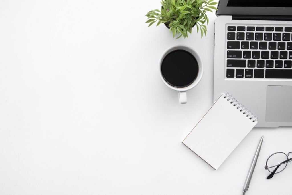 A flat lay of a laptop, cup of black coffee, small plant, notepad, pen, and a pair of rimmed glasses.