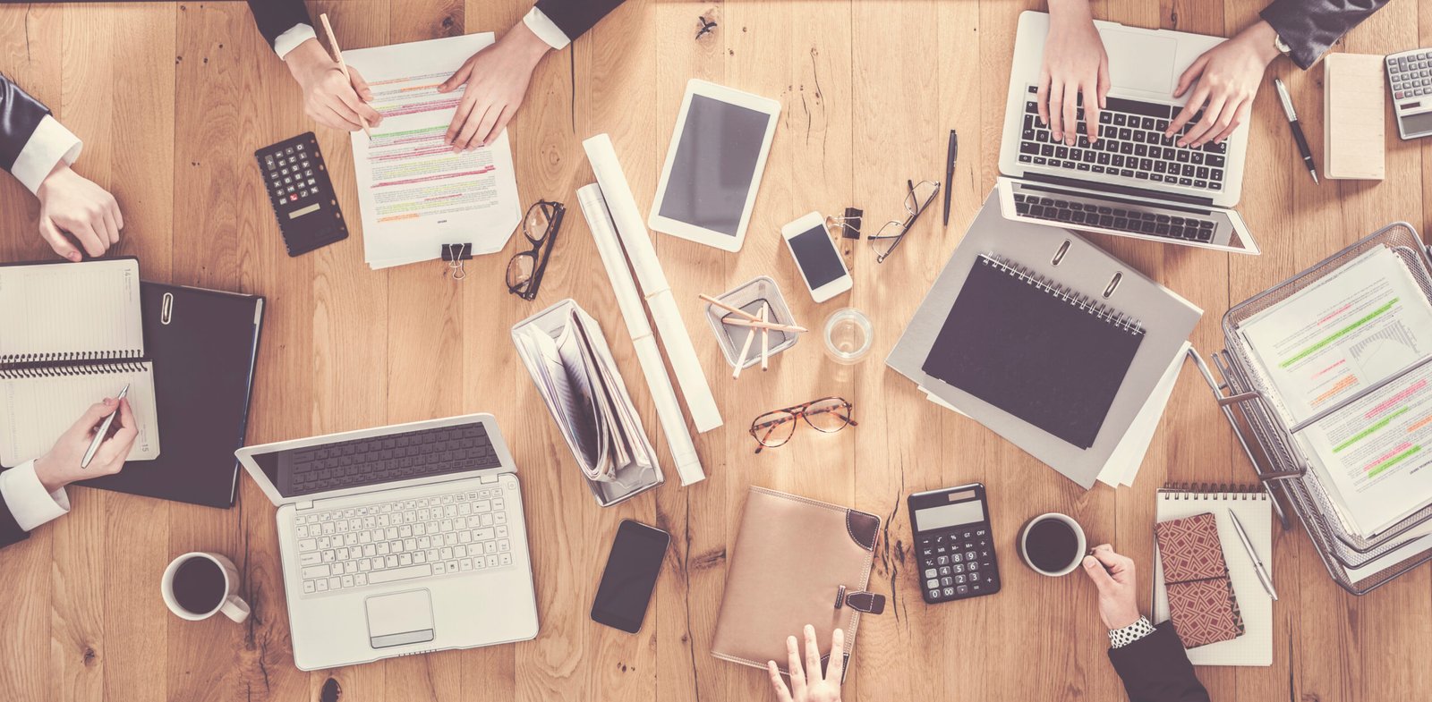 Cluttered wooden desk with the hands of four business people working.